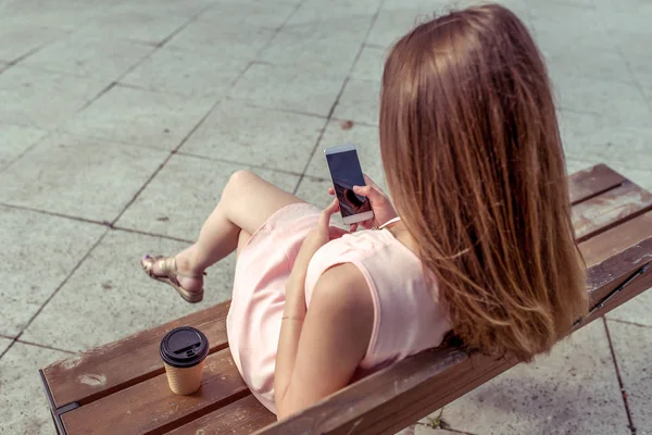 Ragazza in estate in città si siede su sky-bike. Nelle mani del telefono è un'applicazione on-line di reti personali, tazza di tè al caffè. Vestito rosa, vista da dietro, selezione app touch screen con le dita . — Foto Stock