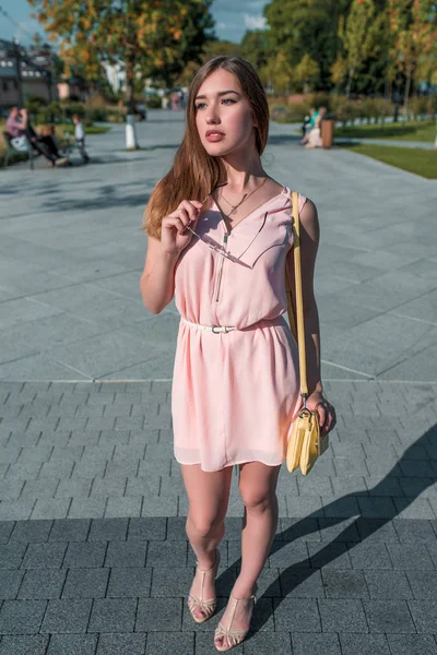 Chica en verano en la ciudad, sandalias de vestir rosa. En manos de gafas de sol. El bolso amarillo, vista desde el frente, abeto verde de fondo, adoquines. Un fin de semana de descanso esperando amigos y novias . — Foto de Stock