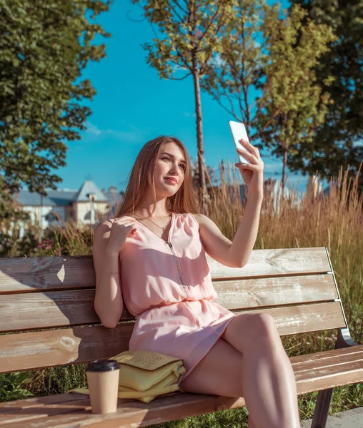 Chica en la ciudad de verano, vestido rosa se sienta en el banco. En su teléfono móvil de la mano, lee escribe el mensaje de Internet, fotografías selfie. Videollamada, grabación, conversación telefónica. Vacaciones de fin de semana en parque . — Foto de Stock