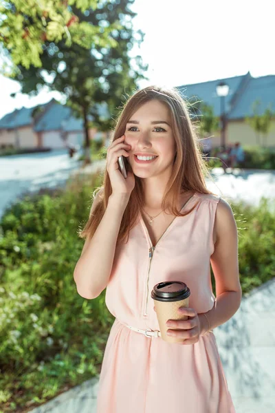 pembe elbiseli güzel kız mutlu gülerek ve neşeli gülümseyen, elinde cep telefonu, kahve çay fincan konuşuyor. Yaz aylarında şehir parkında, parlak güneşli bir günde. Duygular zevk ve zevk.
