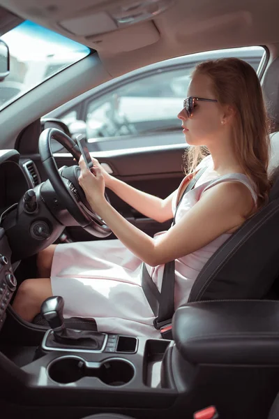 Menina no carro, unidade da direita, tráfego da esquerda, smartphone sua mão, lê escreve mensagem Internet, redes sociais, estacionamento perto do centro comercial. Um longo engarrafamento, paragem forçada, longa espera . — Fotografia de Stock