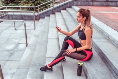 Kadın atlet, merdivenlerde otururken, cep telefonunu elinde tutarak, kulaklıkla müzik dinleme, sabah koşusunda yaz lık egzersiz, fitness motivasyonu genç kadın yaşam tarzı.
