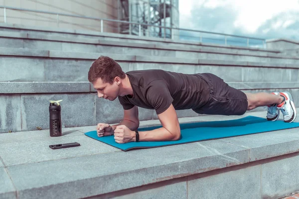 Male athlete, stands in bar, training muscles of back and abs, lifestyle fitness style will rob motivation. In summer in city. Shaker with water and protein, a timer application on a smartphone.