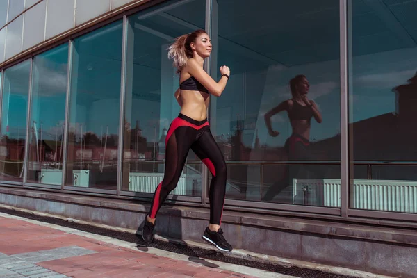 Menina bonita corre no verão na cidade contra o fundo de janelas de vidro do edifício, estilo de vida ativo saudável e elegante, treino de motivação fitness. Espaço livre para texto . — Fotografia de Stock