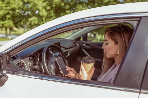 Girl driving car, stands in parking lot, summer waiting for friends and girlfriends, reads message on her smartphone, holds cup of tea her hands with coffee. Summer city background trees. — Stock Photo, Image