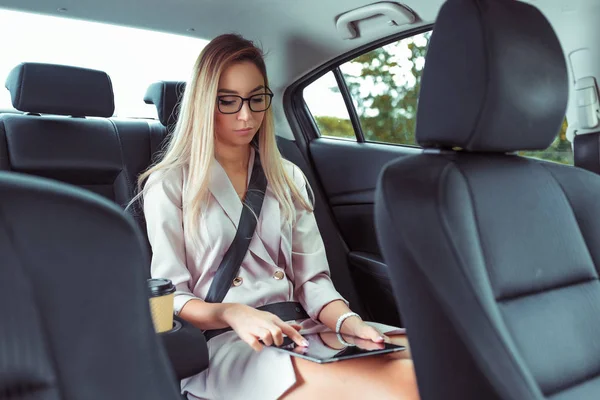 Üzleti nő öltönyt, ül VIP taxi, tartja tabletta a kezében, prések ujját érintőképernyő, csésze kávé tea, a nyári város, gyönyörű nő a szemüveg titkár működik a közúti. — Stock Fotó