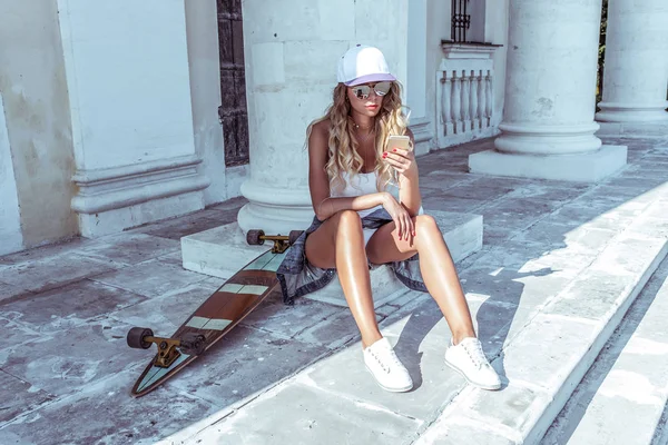 Schöne Frau im Sommer in der Stadt sitzt auf einem Handy, liest eine Nachricht, in den sozialen Netzwerken. Skateboard, Entspannung nach sportlicher Fitness, Workout. braun gebrannte Figur mit langen Haaren. — Stockfoto