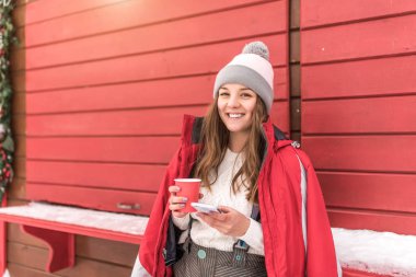 Mutlu kız dükkan kıvrımları arka plan kırmızı çit duruyor, kış sokak hafta sonu şehir. Sıcak ceket şapka, sıcak kahve çayı ile el fincanı, cep telefonu, eğlenmek, dinlenme, gülme, metin için boş alan.