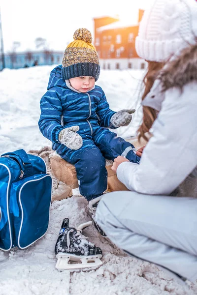 Anak laki-laki berusia 3,5 tahun, musim dingin di arena skating kota, duduk di bangku cadangan, ibu wanita mengganti pakaian anaknya, memakai sepatu, beristirahat pada akhir pekan, salju drift, pakaian hangat, topi, jaket . — Stok Foto