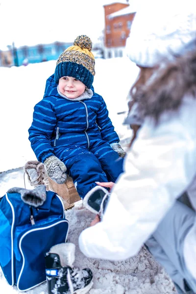 Kisfiú 3,5 év kék overall, télen a város korcsolyapálya, ül egy padon, anya nő cipő gyermek, cipő, korcsolyát, hétvégi ünnepek hó sodródik, meleg ruha kabát kalap. — Stock Fotó