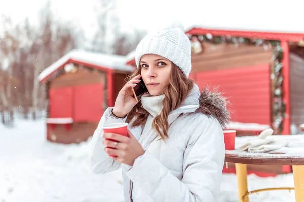 Młoda dziewczyna, Kobieta, rozmowy telefoniczne, w zimie ciepły płaszcz i kapelusz, posiada filiżankę kawy herbaty ręce, rozgrzeje się, słucha wiadomości w Internecie aplikacji internetowych na jej smartphone. — Zdjęcie stockowe