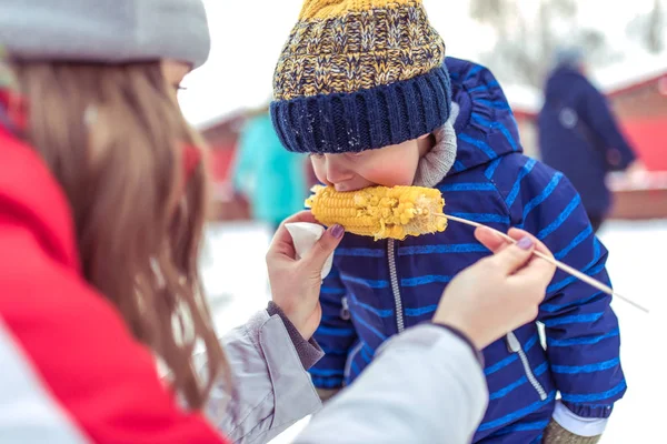Маленький мальчик ест вареную кукурузу на зимней ярмарке, по выходным на природе, синий теплый комбинезон, теплую шляпу, мать кормит ребенка, завтракает и обедает на природе зимой. Курорт с детьми . — стоковое фото