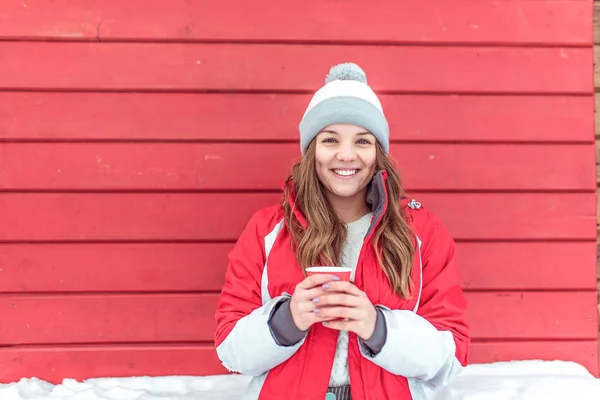 Wanita bahagia tersenyum, berdiri latar belakang selendang toko pagar merah, gadis musim dingin kota akhir pekan jalanan. Topi jaket hangat, tangan dalam cangkir dengan teh kopi panas, bersenang-senang, beristirahat, tertawa, ruang kosong untuk teks . — Stok Foto