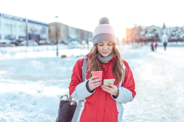 girl winter city background snow snowdrifts, stands warm jacket hat, holds cup tea with hot coffee drinks, writes message smartphone, social networks read online watch video on phone.