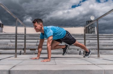 Mavi tişörtlü bir erkek sporcu, yazın şehirde jimnastik yogası yapıyor, sabahları temiz havada egzersiz yapıyor. Gençlik yaşam tarzı, aktif modern erkek atlet, konsept motivasyon hareketi.