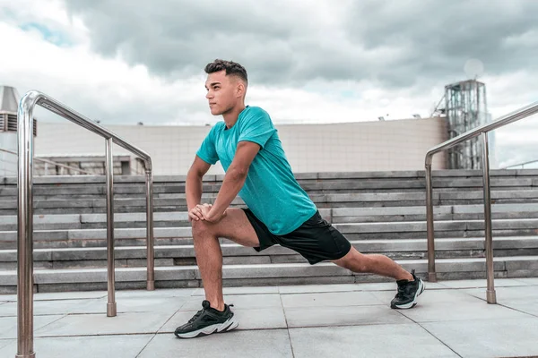 A male athlete in the summer in the city does stretching, preparing for jogging, training in the morning in the fresh air. Youth lifestyle, active modern guy athlete.
