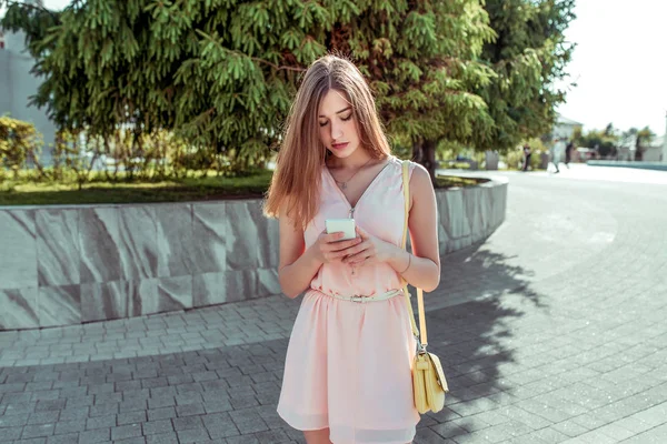 Een studente in de zomer in de stad staat in een roze jurk, schrijft een bericht over de telefoon, leest nieuws op sociale netwerken op internet, vrije ruimte voor tekst, een gele handtas, achtergrond bomen, gazon. — Stockfoto
