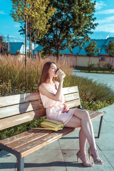 Mädchen in rosa Kleid im Sommer in der Stadt, sitzt auf Bank, trinkt Tee Kaffee, Hintergrund Park Bäume, gelbe Handtasche, Pause Snack, Mittagessen nach der Arbeit im Park. — Stockfoto