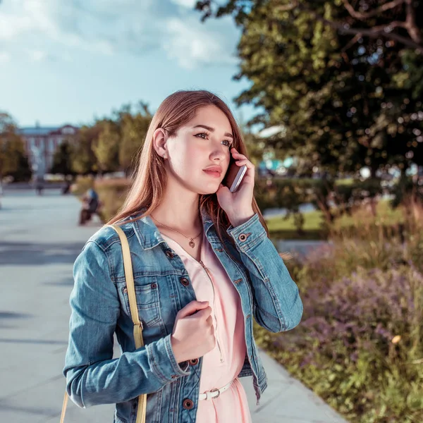 Gadis di musim panas di taman kota, memanggil ponselnya, dalam blus merah muda dan jaket denim, rambut panjang, riasan santai. Emosi kenyamanan dan perhatian mendengarkan lawan bicara . — Stok Foto