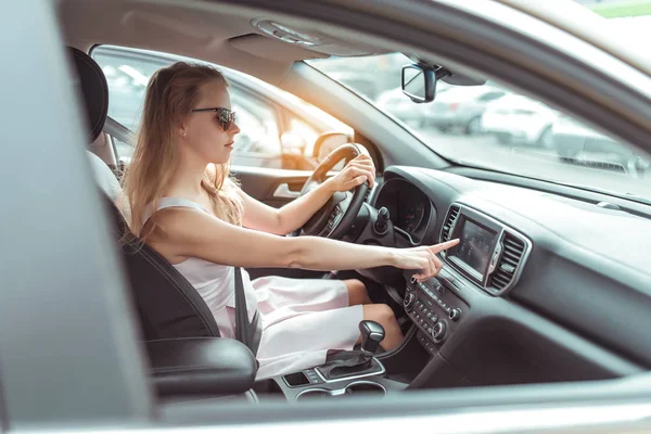 Egy nő az autóban, navigációs alkalmazást választ érintőképernyős kijelzőn. rózsaszín ruha. automata sebességváltó, parkolóban a bevásárlóközpont mellett, autós belső tér, rádióállomás választás. — Stock Fotó