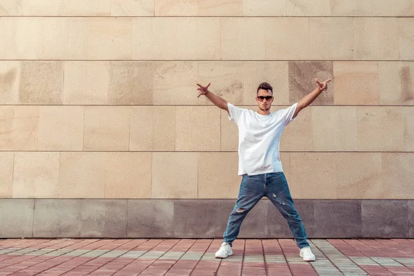 young guy a dancer in a white T-shirt, jeans, dancing break dance in summer in the city, in sunglasses, white sneakers background building clouds, active hip hop, youth lifestyle