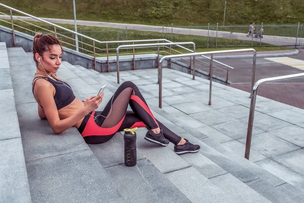 Vakker jente i sportstøy, om sommeren i byen hviler hun etter hard trening i frisk luft, lytter til musikk på hodetelefoner, ser på videoppodcast, fritt rom for motivasjonstekst . – stockfoto