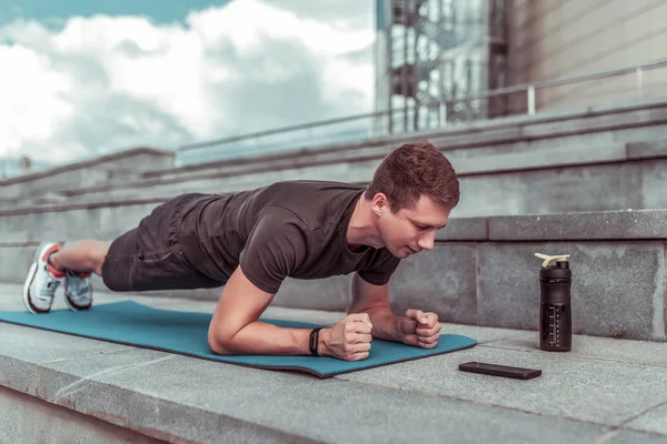 夏の街でトレーニング中の男は横に強調スタンド,板,腹筋トレーニング,ストレッチ柔軟性,スマートフォンのアプリケーションの電話タイマーに見えます,タンパク質と水とシェーカー. — ストック写真