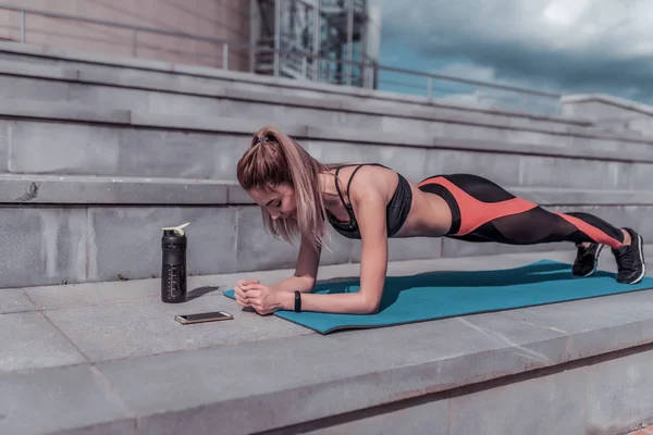 Girl training summer in city, stands emphasis lying down, plank, abs training, abdominal muscles woman back stretching flexibility, smartphone application phone timer, shaker with protein water.