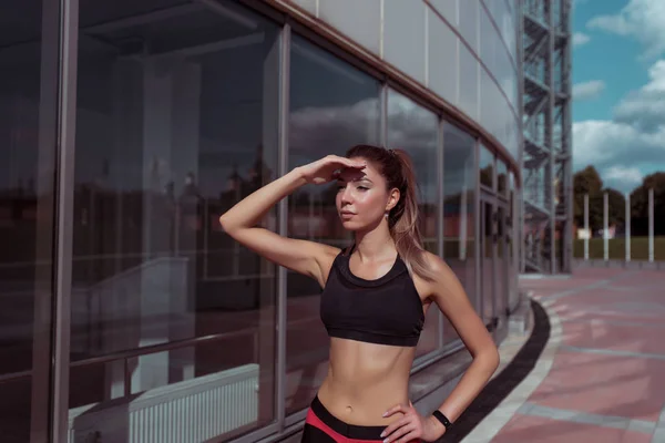 Bela menina bronzeada atlética olha para a distância, cobre a mão do sol, no fundo da cidade de verão de janelas de vidro, espaço livre para texto motivação fitness. Top Sportswear . — Fotografia de Stock