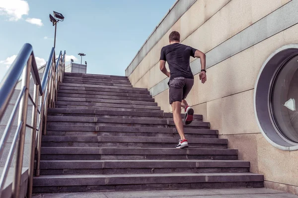 Athlete man runs in summer in city on steps, morning run, free space for text motivation fitness workout in fresh air. Jumping view from back. Sportswear shorts T-shirt Sneakers.