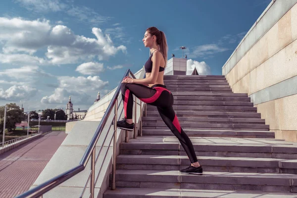 Bonito slim elegante atleta menina, cedo treino matutino faz alongamento músculos antes de correr, espaço livre texto motivação fitness treino. Sportswear leggings topo, céu fundo escadas edifícios . — Fotografia de Stock