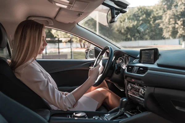 Girl driving car in summer city, in parking lot in traffic jam while waiting, looks phone, reads and writes message in smartphone. Long hair formal suit business lady. Interior car automatic gearbox. — Stock Photo, Image