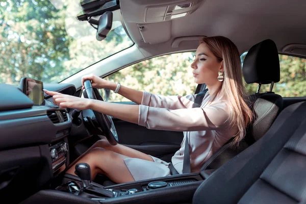 woman in pink suit in car, behind right wheel of car, selects application on touch screen display, selects route, in summer in city, business lady goes to meeting.
