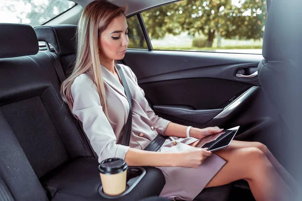 Hermosa mujer de negocios paseos en coche de pasajeros compartimiento de pasajeros en taxi VIP, sujeta la tableta en sus manos, lee y ve video. Cabello largo, piel bronceada, taza de café bebida de té . — Foto de Stock