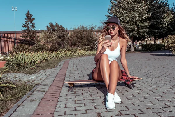 Girl summer city, sits on board for skateboard riding longboard, reads and writes message mobile phone, free space for text. Resting watching video on display screen. Tanned figure beautiful woman. — Stock Photo, Image