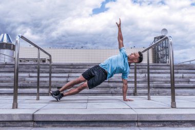 Erkek sporcu, yaz tatilinde şehirde yoga yapıyor. Isınma hareketleri yapıyor. Kol kola bir bar, sabah koşusundan önce spor, bulutun arka planında somut adımlar..