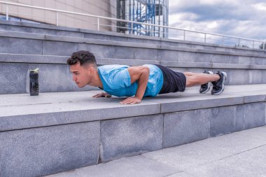 Erkek sporcu, bronzlaşmış adam, yaz şehri, şınav, yedek kulübesinde uzanmak, göğüs eğitimi, pres barı, su dolu şişe proteini, fitness egzersizi, arka plan somut adımlar. Motivasyon metni için boş alan.
