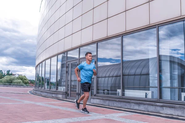 Athlete man runs in summer in city on a morning run, background glass windows of building, free space for text motivation. Active youth lifestyle, fitness workout man.