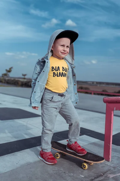 3-4岁的小男孩，秋天的夏天在城市里，滑板训练，开心的微笑，休闲装牛仔夹克，棒球帽。 那男孩骑在木板上. — 图库照片