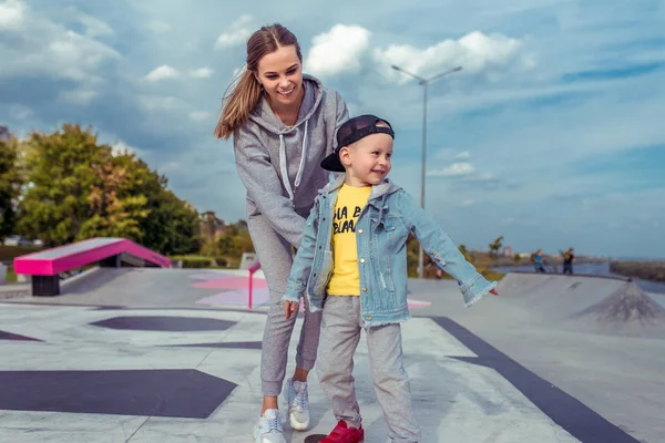 Ibu yang bahagia bermain dengan anak laki-lakinya yang berusia 3-5 tahun, di musim gugur di kota, pelatihan skateboard, bahagia tersenyum, pelatihan. Anak itu naik di papan . — Stok Foto