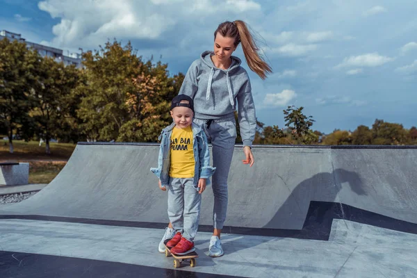 महिला माँ अपने बच्चे के साथ 3-5 साल का एक छोटा लड़का खेलती है, शहर में शरद ऋतु की गर्मियों में, स्केटबोर्ड प्रशिक्षण, खुश मुस्कुराते हुए, प्रशिक्षण प्रशिक्षण। लड़का बोर्ड पर सवारी कर रहा है . — स्टॉक फ़ोटो, इमेज