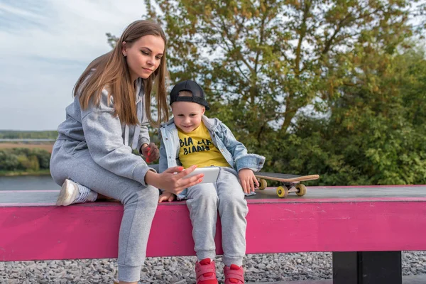 Young mother with a boy 3-5 years old son, watching a video on the phone, video call, summer autumn day in a city park, skateboard, casual clothes fun, joy laughter. — Stok fotoğraf