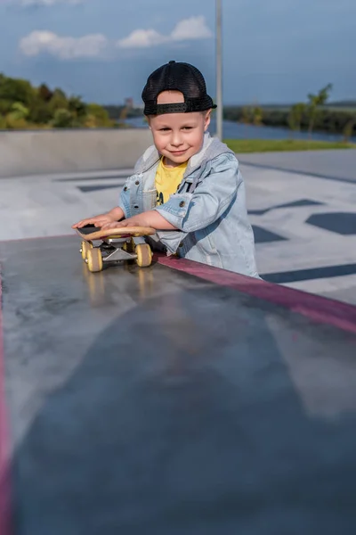 Anak laki-laki berusia 3,5 tahun, anak tersenyum, bermain, telah beristirahat, pada hari musim panas di taman kota, menyenangkan pakaian santai, papan skateboard di tangannya, pakaian santai . — Stok Foto