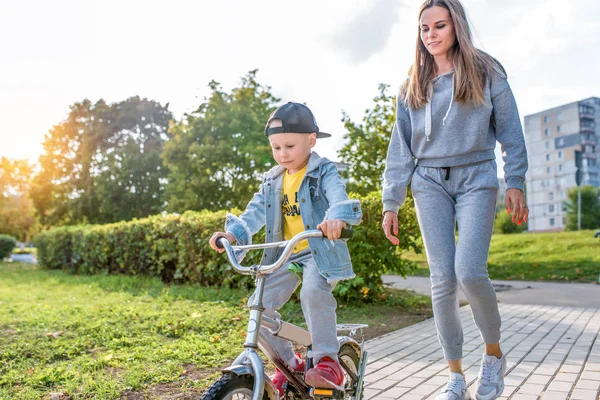 Mama tanara antreneaza copil, baietel de 3-5 ani, copilul invata primele lectii de bicicleta, echilibru, odihna, o zi de toamna de vara in parcul orasului, hainele casual sunt distractive . — Fotografie, imagine de stoc