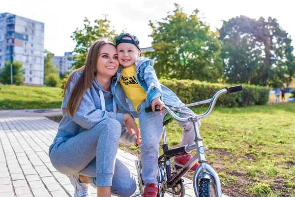 Young happy family smiling happy rejoicing, mom trains child little boy 3-5 years old child, learns first lessons of bicycle, balance, relax, summer autumn day in city park, casual clothes fun. — Stok fotoğraf