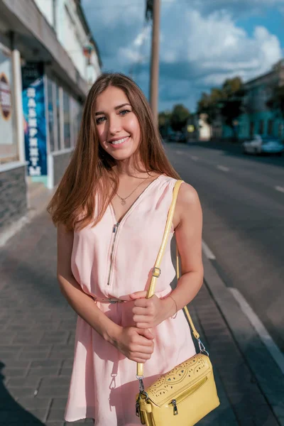 Beautiful and happy girl stands in summer city, smiles, road is background, yellow handbag hands, she is waiting for friends and girlfriends, long hair tanned slim figure. Розовое платье случайный макияж . — стоковое фото