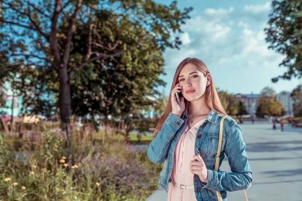 girl student summer city stands autumn spring, pink dress, smartphone hand, makes phone call, listens message online app Internet, park trees background. Happy smiling joyful resting. Free space text.