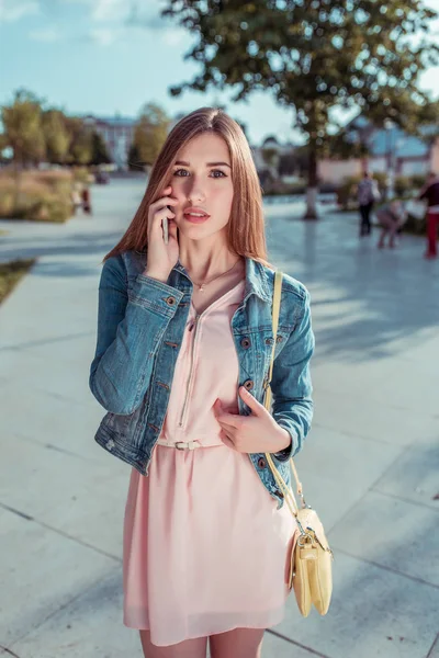 Красивая девушка студентка стоя летом города, руки смартфона звонки слушает сообщение, желтая сумка, ожидая друзей подруг, длинные волосы, загорелая стройная фигура. Розовое платье случайный макияж . — стоковое фото