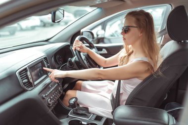 girl in car selects application on touch screen, navigation application on Internet, strapped holds wheel, left-hand traffic, wheel on right side. Woman in summer sunglasses in city. clipart