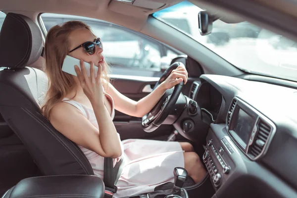 girl in cars interior calls on phone, looks in rearview mirror, parks at parking lot of shopping center, holds it fastened by steering wheel. Woman in summer sunglasses in city wearing a pink dress.